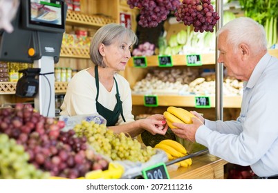 Mature Woman Cashier In Apron Standing At Counter In Greengrocer. Senior Man Purchasing Bunch Of Bananas.