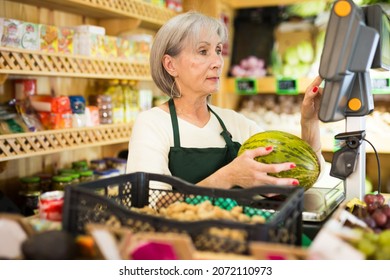 Mature Woman Cashier Weighting Melon While Standing At Counter In Greengrocer.