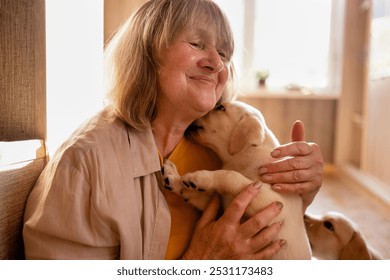 A mature woman in casual clothes hugs a puppy at home. A pet licks its owner face. A pensioner takes care of a small golden retriever. A middle-aged woman smiles and caresses a cute dog - Powered by Shutterstock