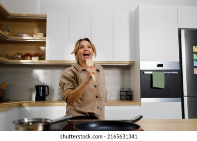 Mature Woman Cooks Food In The Kitchen And Sings Using A Wooden Spatula As A Microphone