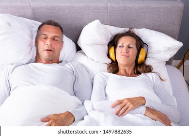 Mature Woman Covering Her Ears With Headphone While Man Snoring In Bed - Powered by Shutterstock