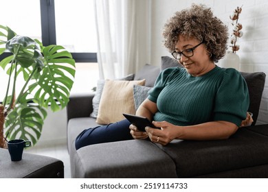 A mature woman with curly gray hair and red glasses sits on a sofa, using a tablet. She is focused and engaged, dressed casually in a green sweater, in a comfortable home setting - Powered by Shutterstock