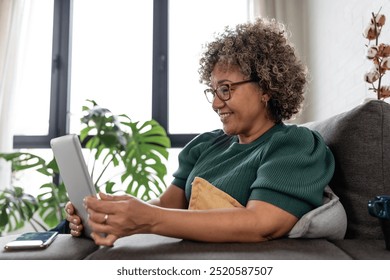 A mature woman with curly gray hair and red glasses sits on a sofa, using a tablet. She is focused and engaged, dressed casually in a green sweater, in a comfortable home setting - Powered by Shutterstock