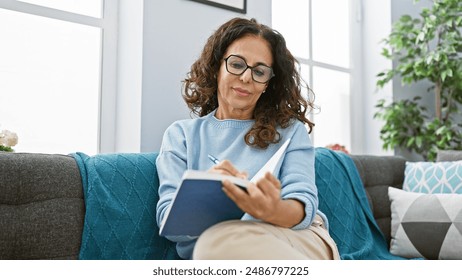 Mature woman with curly hair writing in a notebook while sitting on a blue sofa indoors. - Powered by Shutterstock