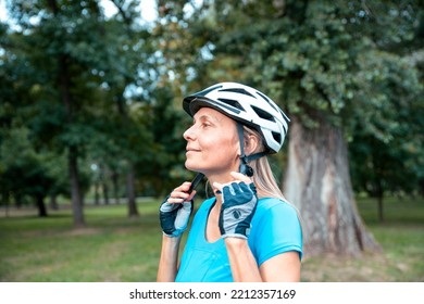 Mature Woman Cyclist In Sports Clothes Puts On Head Cycling Helmet. Preparing For The Ride In The Green Forest Park. Copy Space