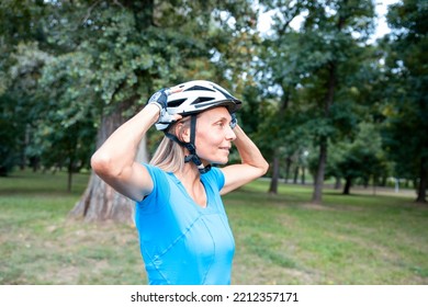 Mature Woman Cyclist In Sports Clothes Puts On Head Cycling Helmet. Preparing For The Ride In The Green Forest Park. Copy Space