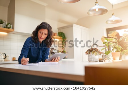 Mature Woman With Digital Tablet Reviewing Domestic Finances And Paperwork In Kitchen At Home