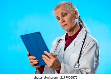 Mature Woman Doctor Medical Service Health Care Physician Looking At Tablet Computer Isolated On Solid Blue Background Copy Space.