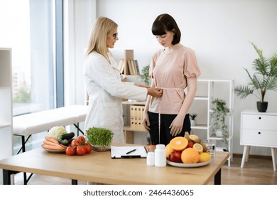 Mature woman in doctor's coat finding out waist length of female patient with measuring tape in consulting room. Efficient dietitian calculating body mass index for assessing healthy weight. - Powered by Shutterstock