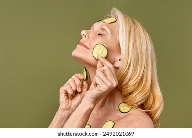 Mature woman doing spa treatments, rejuvenating her facial skin with cucumber slices for a refreshing and healthy skin care isolated on an olive background. Cosmetology and age skin care. - Powered by Shutterstock