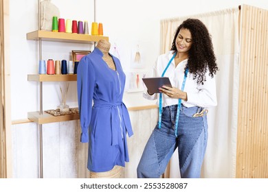 A mature woman, dressed casually, reviews her fashion design ideas on a tablet. She stands beside an elegant dress and colorful spools, showcasing her creative workspace. - Powered by Shutterstock