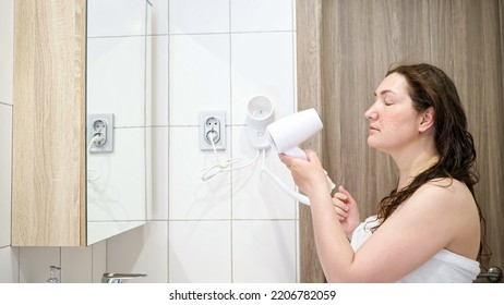 Mature Woman Dries Wet Hair With Hairdryer And Looks In Mirror. Lady Wrapped In White Towel During Usual Morning Routine After Taking Shower