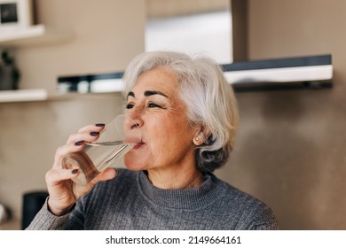 Mature woman drinking fresh clean water from a glass. Grey-haired senior woman staying healthy and hydrated at home.