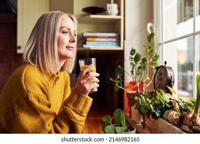 Mature Woman Drinking Glass Of Water In The Kitchen Looking Through The Window