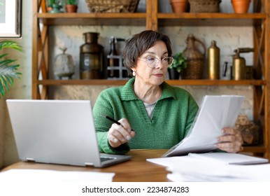 A mature woman during a document check. She checks the correctness of filling out the documentation and enters the data on the site. Modern technology in everyday life concept. - Shutterstock ID 2234874283