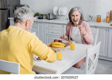 Mature Woman Eating Pancakes During Breakfast With Husband In Kitchen