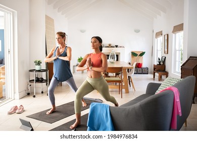 Mature Woman Friends Practicing Yoga At Home. Sporty Fit African Woman Practices Yoga Asana Warrior Pose At Home With Her Friend. Mid Adult Lady Practicing Watching Online Sport Lesson On Tablet.