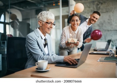 Mature woman getting Birthday surprise from her colleagues while working on a computer in the office. - Powered by Shutterstock