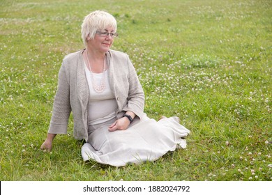 Mature woman in glasses with short blond hair in linen clothes
  sitting on a grass in summer park