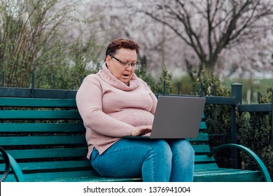 Mature Woman In Glasses Using Laptop, Is Scared, A Saw Which Also Should Not See, Dressed In A Pink Sweater And In Jeans Sitting On A Chair In The Park