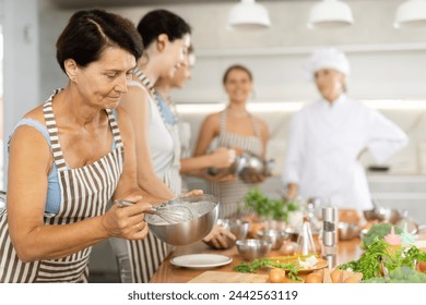 Mature woman at group cooking class holds bowl in hands and mixture cream sauce with white wine for meat with whisk - Powered by Shutterstock