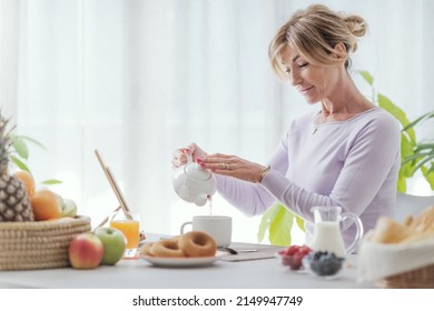 Mature woman having breakfast at home, she is sitting and pouring tea in a cup - Powered by Shutterstock