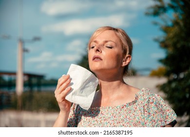 Mature Woman Having Hot Flashes Outdoors, Trying To Cool Herself With Paper Tissue. Blonde Female Portrait Cooling Down On Hot Summer Day