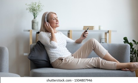 Mature woman in headphones enjoying music, using phone, peaceful satisfied older female with closed eyes relaxing on comfortable couch at home, listening to favorite song, leisure concept - Powered by Shutterstock