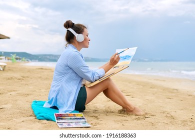 Mature Woman In Headphones Sitting On Sandy Beach Drawing Sketch Of Sea Nature With Watercolors. Rest, Relaxation, Hobbies And Leisure, Creativity, Mental Health, Middle-aged People