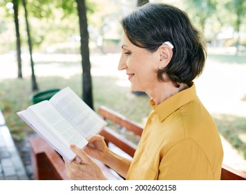 Mature Woman With A Hearing Impairment Uses A Hearing Aid In Everyday Life, Reading A Book In Park, Outdoor. Hearing Solutions