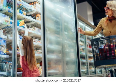 Mature Woman And Her Grandchild Shopping At Supermarket, Taking Product From Fridge