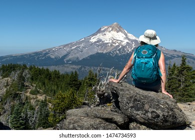 A Mature Woman Hiker Sites On A Rock With Her Back To Us And Enjoys The View Of Mt. Hood From Lookout Mt. There Is A Clear Blue Sky And Bright Summer Sunlight.