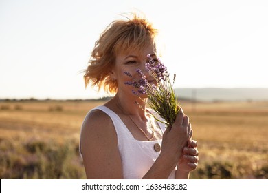 Mature woman holding a bouquette of lavender near face andbreathes in the scent of flowers. Blond slim female dressed white sundress and posing on the field in sunny day