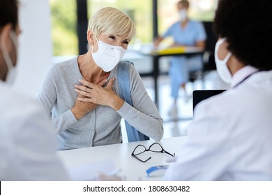 Mature Woman Holding Her Chest In Pain While Wearing Protective Face Mask And Communicating With A Doctor During Medical Appointment At Clinic. 