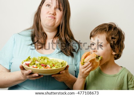 mature woman holding salad and little cute boy with hamburger teasing close up, family food