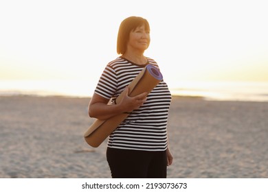 Mature Woman Holding A Sports Mat And Preparing To Practice Yoga Outdoors On Sea Beach. Happy Mature Overweight Woman Exercising On Seashore. Copy Space. Meditation, Yoga And Relaxation Concept