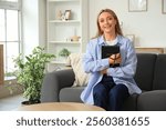 Mature woman with Holy Bible and prayer beads sitting on sofa at home