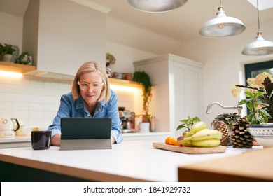 Mature Woman At Home In Kitchen Drinking Coffee And Looking At Digital Tablet