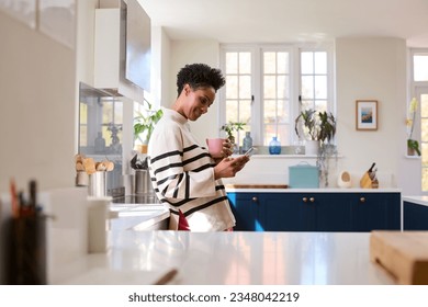 Mature Woman At Home In Kitchen Drinking Coffee And Checking Social Media On Mobile Phone - Powered by Shutterstock