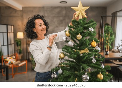 A mature woman joyfully decorates a Christmas tree with sparkling gold and silver ornaments, placing a shining star atop, creating a festive and warm atmosphere at home - Powered by Shutterstock