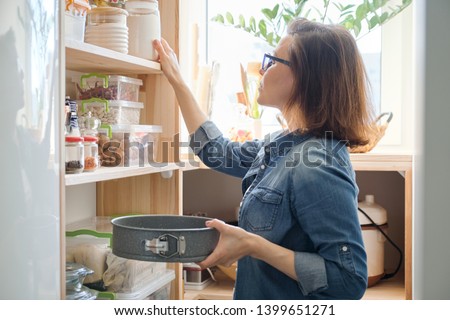 Mature woman in the kitchen pantry with products. Storage wooden stand with kitchenware, products necessary to cook.