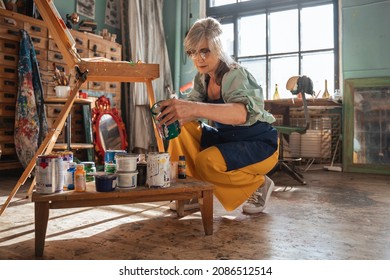 Mature Woman Kneeling And Examining Paints While Painting In Her Home Studio