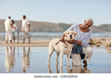 Mature Woman Kneeling Next To A Dog On A Beach At Low Tide.