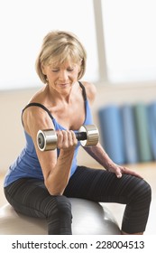 Mature Woman Lifting Dumbbell While Sitting On Fitness Ball At Home