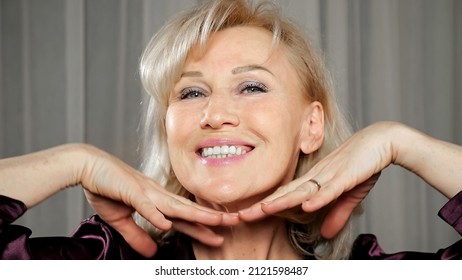 Mature Woman With Long Blonde Hair And Light Makeup Showcases Beauty Of Face And Good State Of Skin Smiling Happily Against Curtain Extreme Closeup.