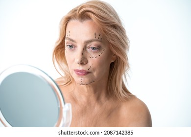 Mature woman looking at the mirror with drawing surgical lines on her face - Powered by Shutterstock