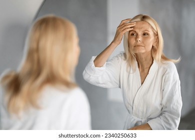 Mature Woman Looking In Mirror And Touching Wrinkles On Her Face, Upset Beautiful Senior Female Examining Fine Lines On Forehead, Suffering Skin Aging, Selective Focus On Reflection, Closeup - Powered by Shutterstock