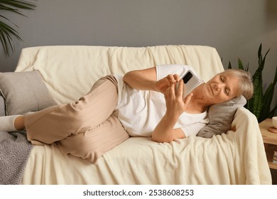 Mature woman lying on sofa at home scrolling through smartphone happily browsing online shopping and checking social media using modern technology and enjoying her retired life - Powered by Shutterstock