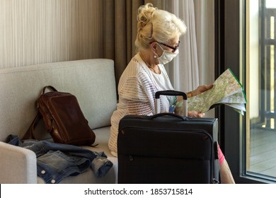 A Mature Woman In A Medical Mask, Reading A Map, Planning A Trip While Sitting In A Room. Black Suitcase In The Foreground. Travel, Tourism During Coronavirus Epidemic. Covid 19.