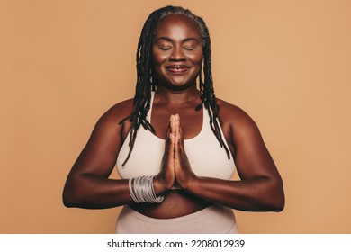 Mature woman meditating with her eyes closed and her hands in prayer position. Black woman with dreadlocks practicing yoga in a studio. Happy middle-aged woman maintaining a healthy lifestyle.